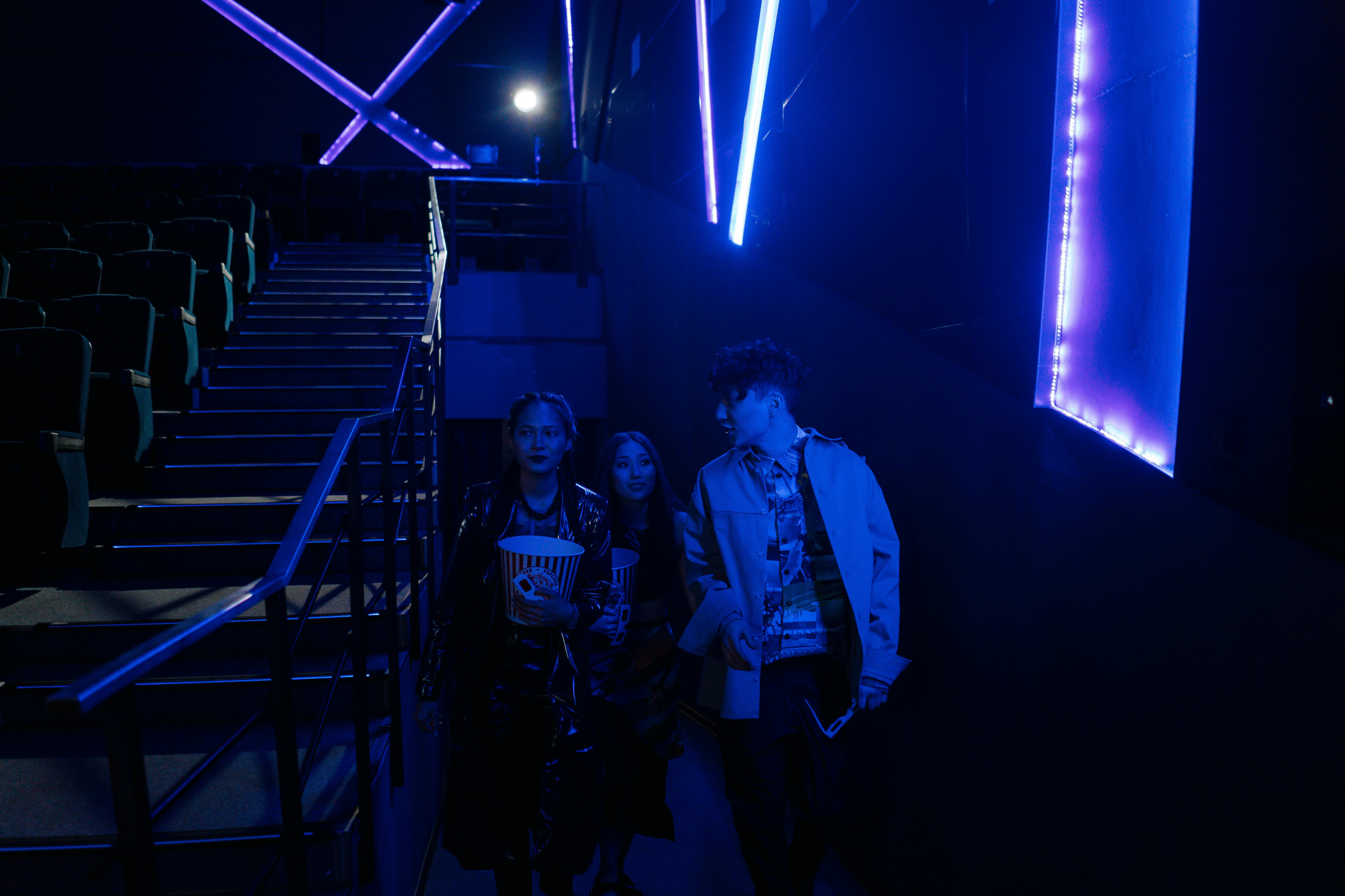 Young adults entering a dimly lit movie theater, holding popcorn with neon blue lighting.