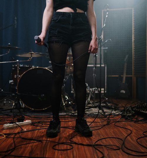 A woman in denim shorts holds a microphone on a wooden floor, surrounded by band equipment.