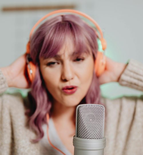 Woman with purple hair singing into a microphone wearing headphones indoors.